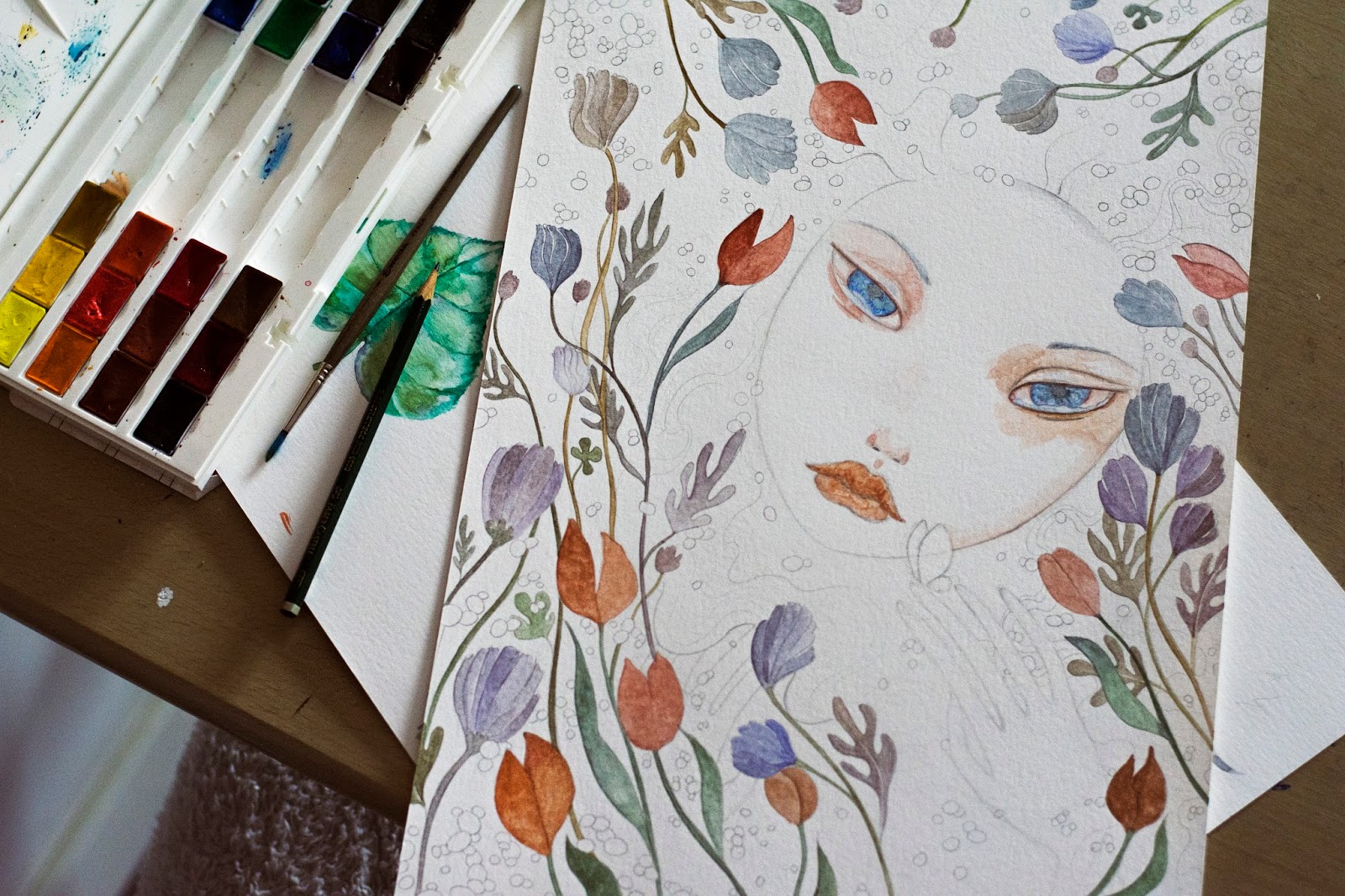 Miniatura de ilustración en proceso: rostro femenino rodeado de flores, paleta y pincel sobre mesa de madera